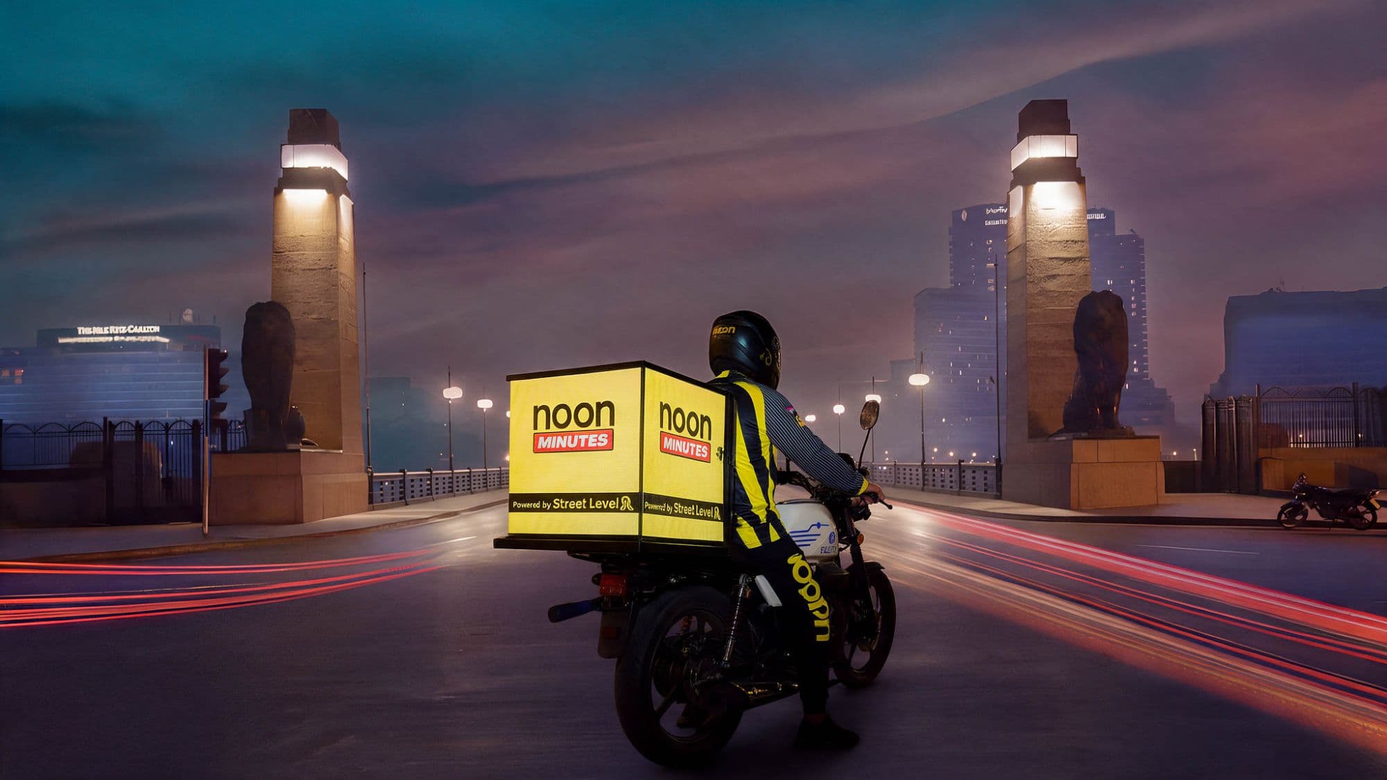 noon Minutes rider on Qasr El Nil Bridge at dusk, with a Street Level screen box on the back of the bike