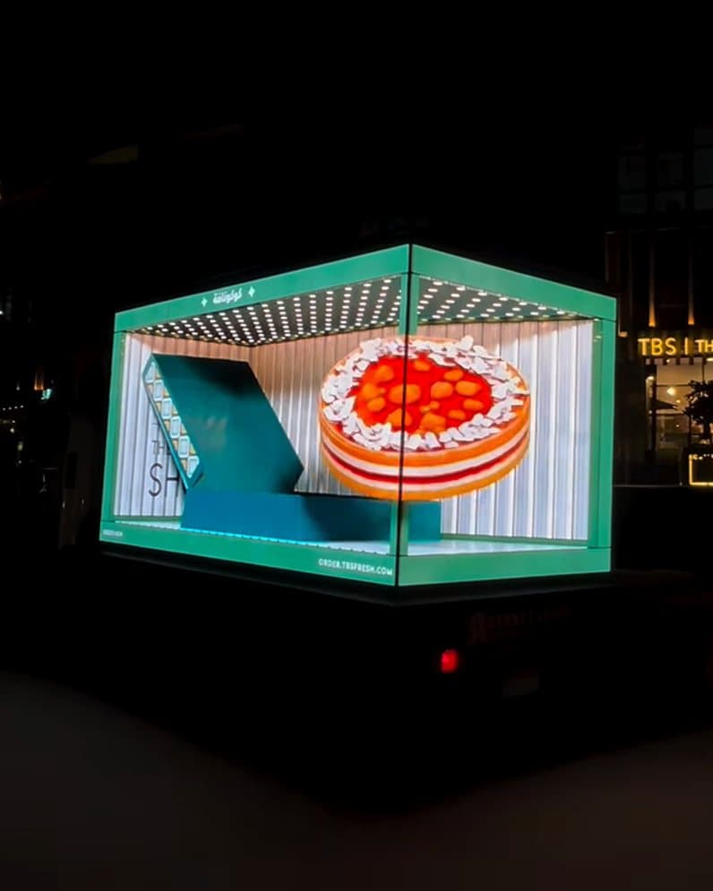 Street Level truck at night showing a 3D TBS cake ad, outside a TBS bakery